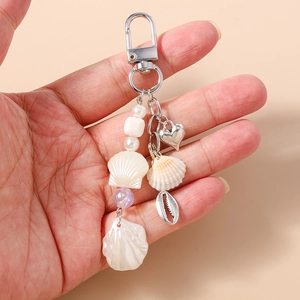 Detail of beaded keychain featuring heart, shells and pearls held by hand on beige background