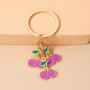 Dettaglio portachiavi ciliegia viola con finitura dorata e foglie verdi su sfondo beige