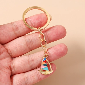 Detail of a gold sailboat keychain with vibrant enamel sails held in hand against light beige