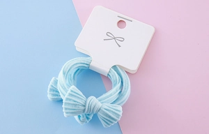 Macro detail of a soft light blue bow hair tie on a white display card over a pastel background