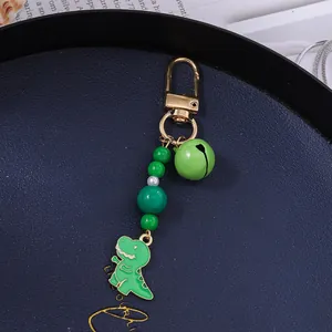 Close-up of a green dinosaur charm with beads and a metallic bell on a gold keychain