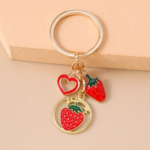 Detail of gold keychain with red enamel strawberry charms and heart pendant on beige props