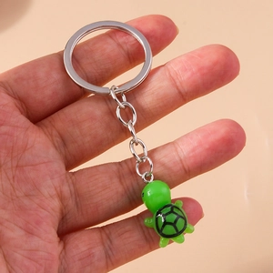 Cute green cartoon turtle keychain with metal ring shown held in hand on neutral background
