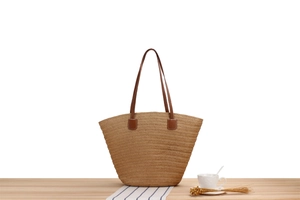 Detail of woven straw tote bag with brown leather straps on a light wooden table surface