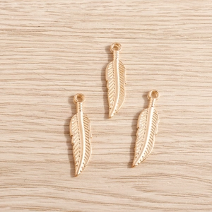 Macro detail of three golden alloy feather charms on a light wood background for DIY jewelry