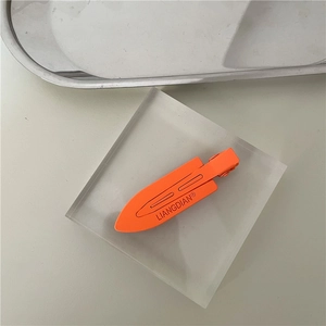 Close-up of bright orange matte metal hair clip with logo on acrylic stand