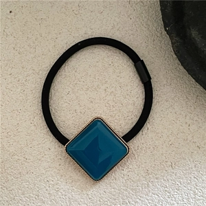 Detailed view of square peacock blue acetate hair tie with golden metallic trim and black band