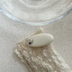 Détail d'une pince à cheveux ovale crème avec cœur argenté sur un tissu en maille