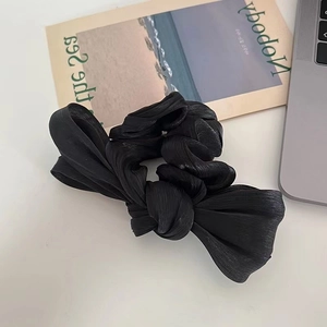 Detail of a large black pleated satin bow scrunchie with a glossy texture on a white surface
