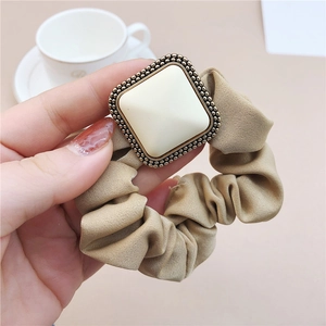 Elegant khaki satin scrunchie with a vintage gold square pendant held against a white cup