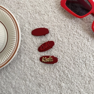 Close-up of three red hair clips with Rich letter design on a textured fabric background