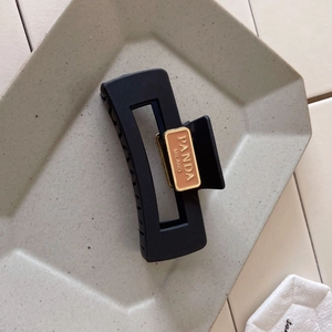 Detail of matte black rectangular hair claw clip with metallic logo plate on gray ceramic