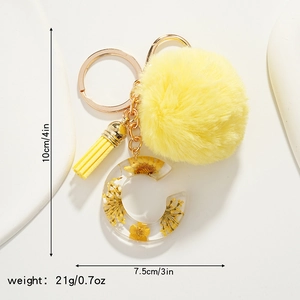 Close-up of yellow resin letter C keychain with dried daisy, fur pompom and tassel