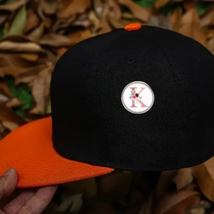 Detail of baseball cap showing black crown and orange brim with embroidered letter K