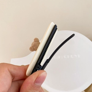 Handheld view showing a plush brown bear hair clip and cream-colored duckbill hair clip
