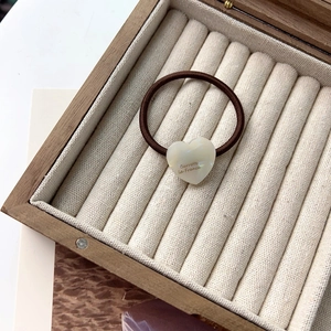 Détail d'un élastique à cheveux cœur blanc perle avec logo doré dans une boîte