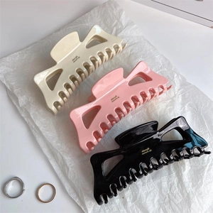 Three oversized acetate hair claws in beige, pink, and black with gold logo on white paper