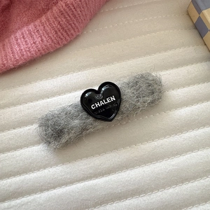 Detail einer grauen Plüsch-Haarklammer mit schwarzem Herz und weißem Logo-Aufdruck