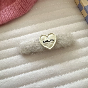 Detail einer grauen flauschigen Herz-Haarklammer mit Herz-Charm auf beigem Polster