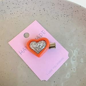 Detail of orange heart hair clip with silver glitter and lucky lettering on a display card