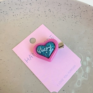 Close-up of heart-shaped hair clip with pink border and blue glitter Lucky inscription