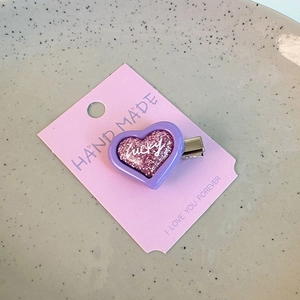 Detail of a purple heart hair clip with pink glitter and white lucky text on a paper card