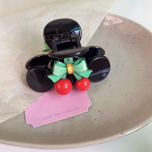 Detail of a black hair claw clip featuring sweet cherry accents and a green bow on a tray