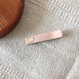 Minimalist pink rectangular hair clip with gold metal label on a beige fabric background