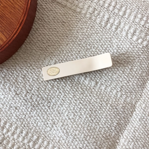 Minimalist matte cream hair clip with oval gold logo tag on soft knit background