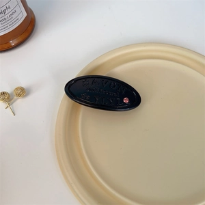 Detail view of a black oval hair clip with embossed soap bar text and a pink rhinestone