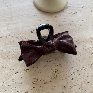 Glossy burgundy leather bow hair claw clip with stitched edges on beige stone surface