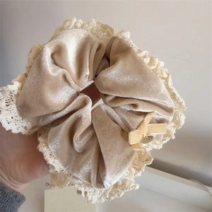 Detail of beige velvet scrunchie with cream lace trim, bow, and gold metal tag