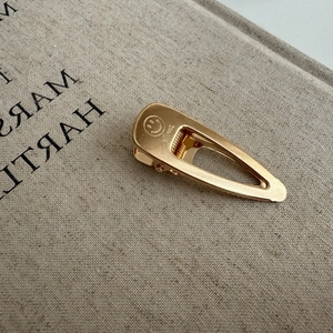 Golden hollow teardrop hair clip with a brushed metal texture and engraved smiley face design