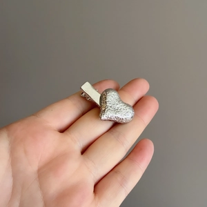Pince à cheveux coeur argenté avec texture martelée tenue à la main sur fond gris uni