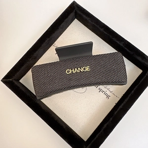Macro detail of a large rectangular black hair claw clip with woven texture and gold metal CHANGE motif