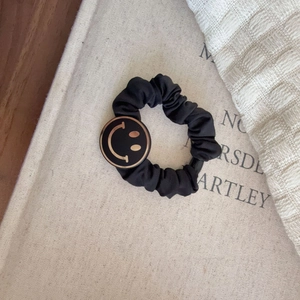 Retro black scrunchie with golden smiley face charm on a textured linen book background
