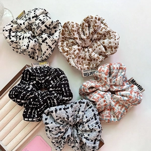 Five assorted tweed checkered scrunchies with brand labels on a beige minimalist setup