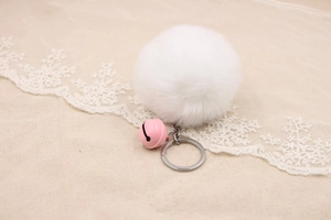 Detail of soft white faux fur pompom keychain with a pink bell on rustic lace fabric