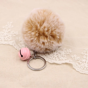 Detail of light brown faux fur pom pom keychain with pink bell on beige lace fabric