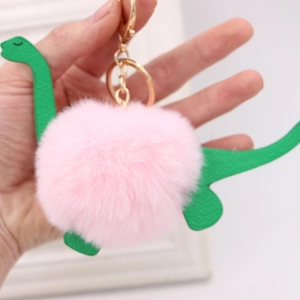 Pink plush dinosaur pompom keychain with green faux leather accents and gold hardware