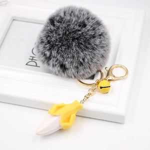 Plush banana keychain with a grey faux fur pom pom and gold bell charm on a white background