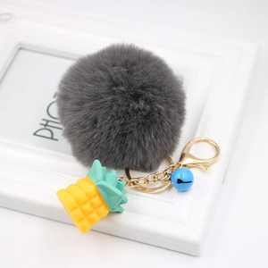 Detail of grey fluffy pompom keychain with yellow pineapple charm and a blue bell