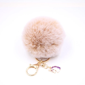 Detail showing soft beige pompom keychain with golden clasp and purple unicorn enamel charm