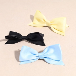 Three elegant satin bow hair clips in yellow, black, and light blue on a cream background