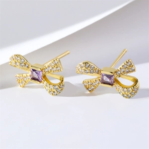 Boucles d'oreilles nœud dorées avec zircon violet et cristaux sur fond de papier blanc