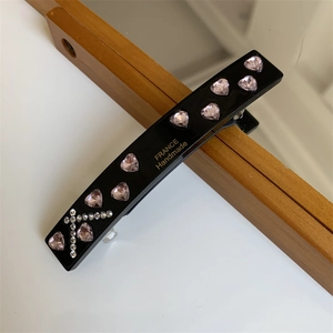 Detail view of black rectangular hair clip with pink heart rhinestones and gold branding