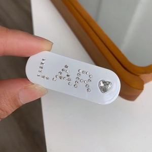 Détail d'une pince à cheveux ovale blanche avec lettrage LA R et strass en forme de cœur