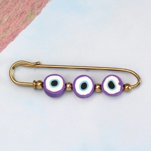 Detail of a golden safety pin brooch featuring three purple evil eye beads and gold accents