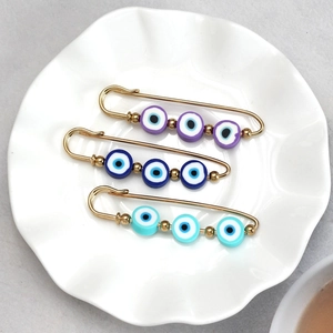 Three gold evil eye safety pins with purple and blue beads in a white ceramic dish
