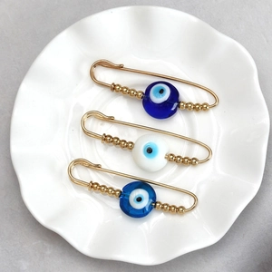 Detailed view of three golden evil eye safety pins with blue beads on a ceramic tray